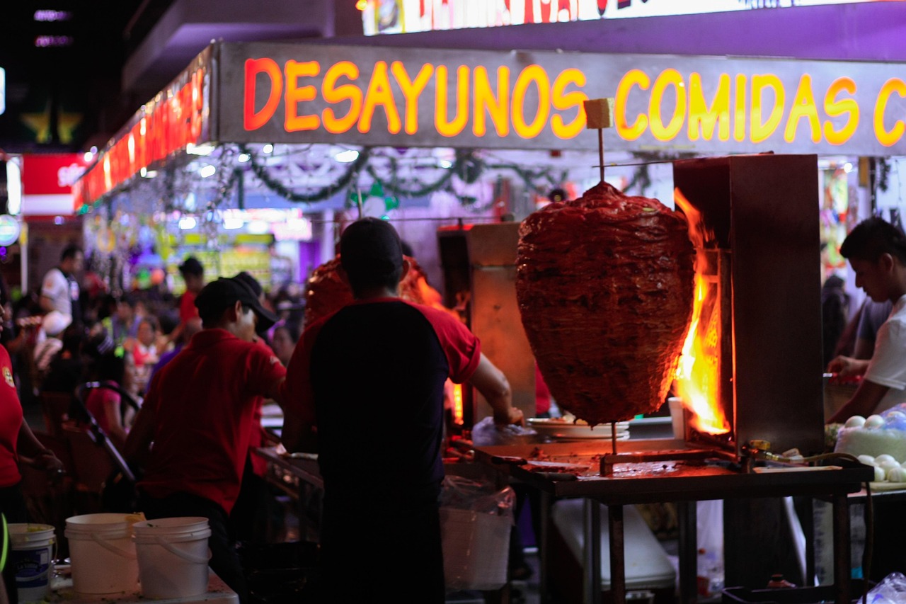 Trompo de tacos al pastor con un gran cono giratorio de cerdo marinado sobre una llama abierta en un concurrido mercado nocturno