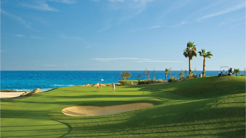 Golfistas en un green junto al mar, con palmeras, búnkeres de arena y el mar de Cortés al fondo
