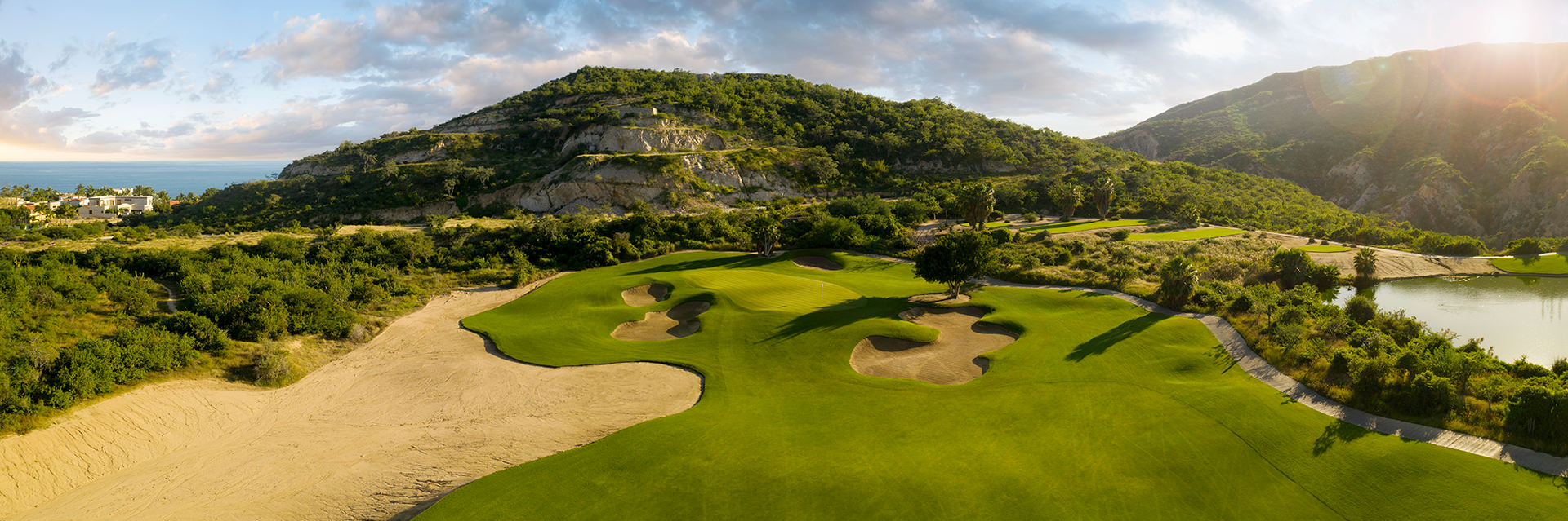 Vista panorámica aérea de un campo de golf con búnkeres de arena, un lago y colinas boscosas bajo un cielo al amanecer