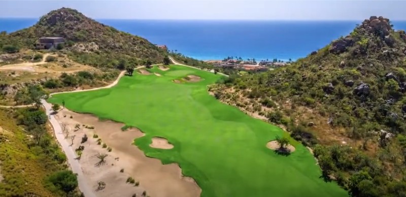 Vista aérea de una calle de golf entre colinas rocosas, con el mar de Cortés al fondo