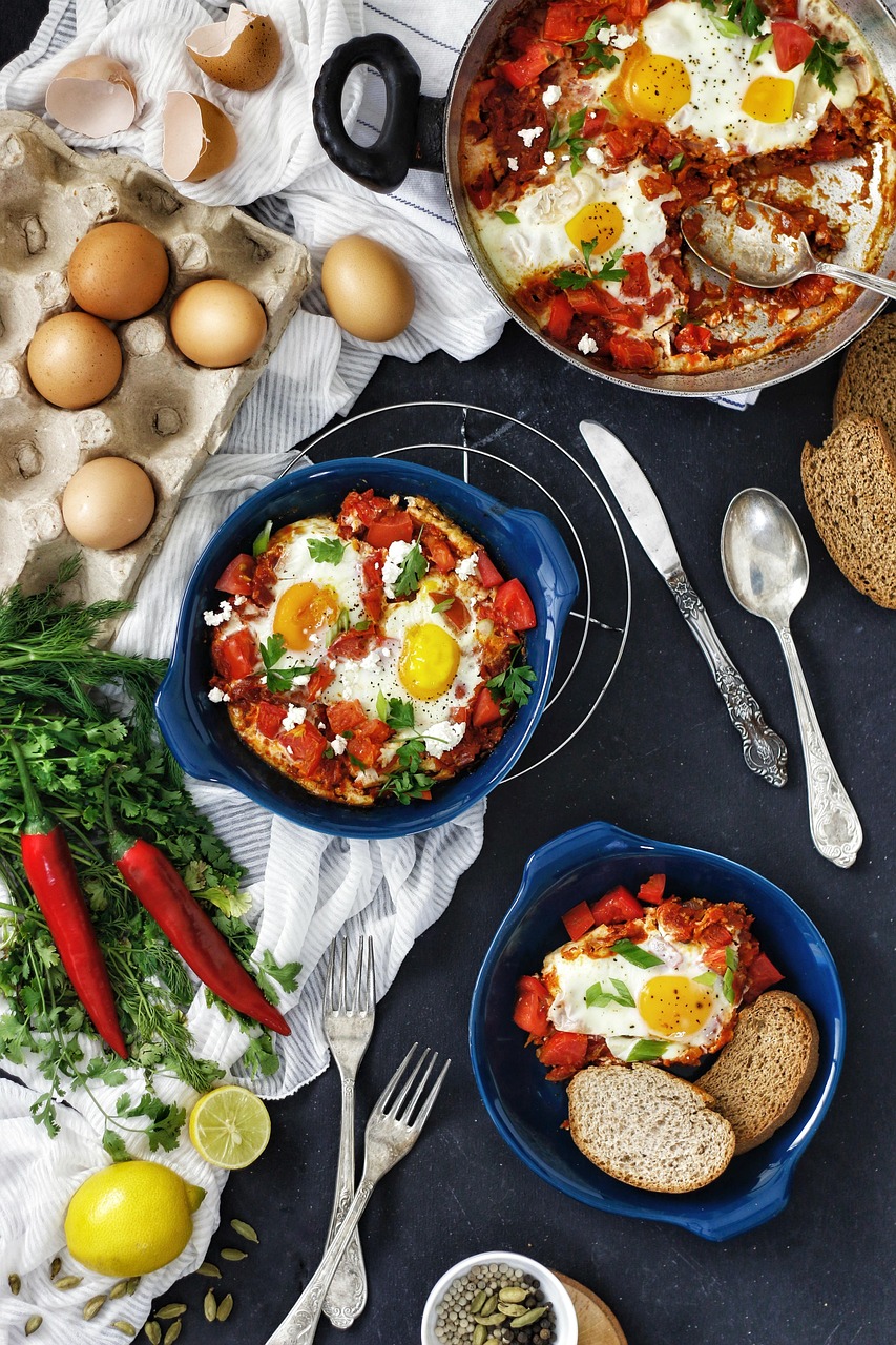 Vista cenital de un shakshuka en una sartén, con huevos en salsa de tomate, pan, hierbas y una bebida de lima sobre una mesa oscura