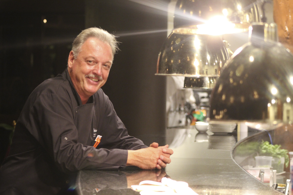 Chef sonriente con chaqueta oscura apoyado en la encimera de una cocina bajo lámparas colgantes