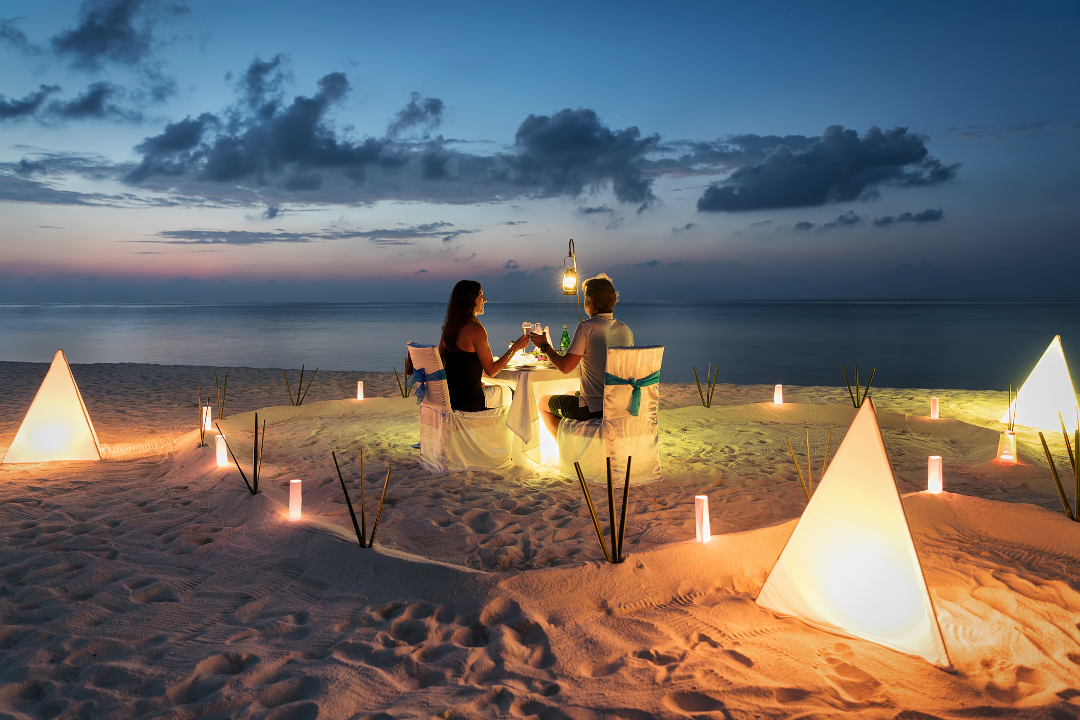 Pareja cenando a la luz de las velas en una mesa en la playa al atardecer, rodeada de faroles piramidales iluminados
