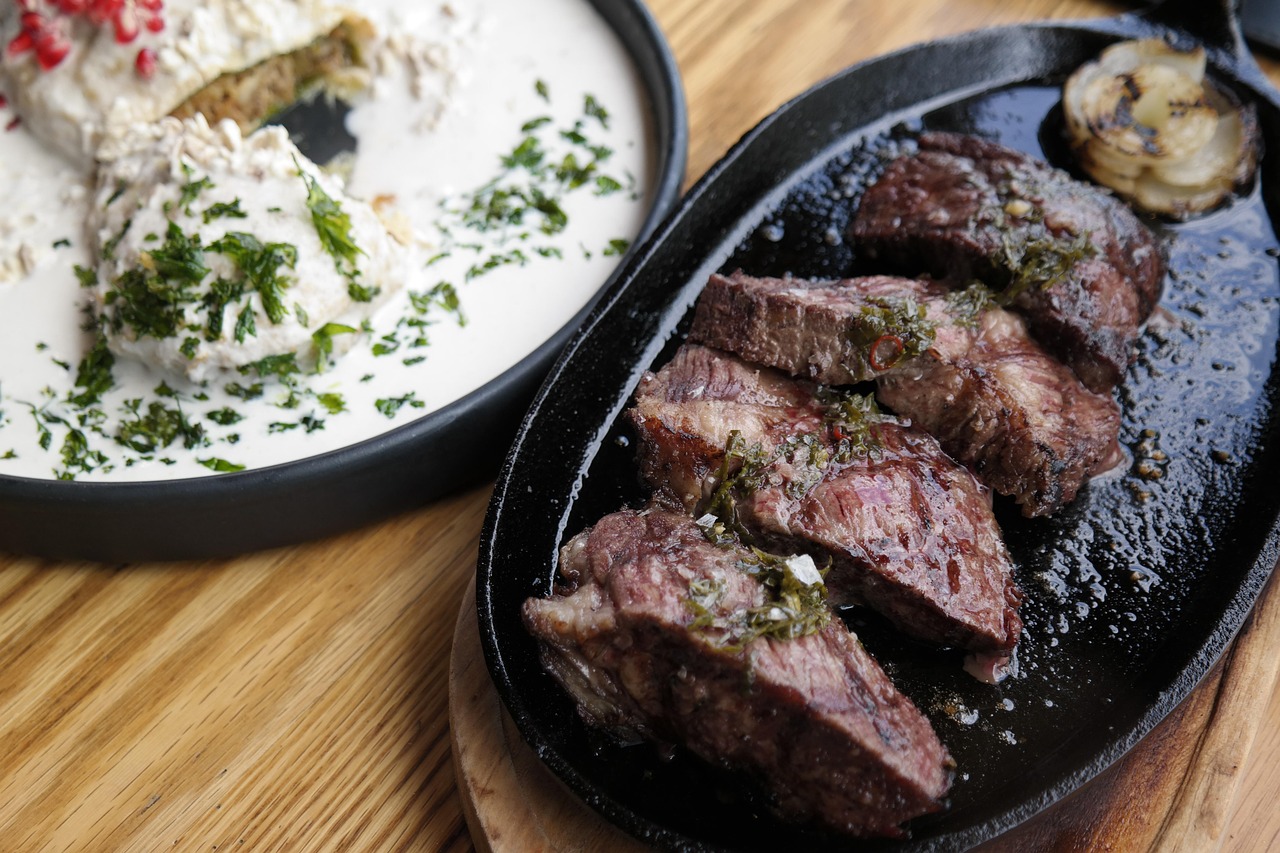 Filete a la parrilla en rebanadas sobre una sartén de hierro fundido, junto a un plato blanco con una salsa cremosa, servido sobre una tabla de madera