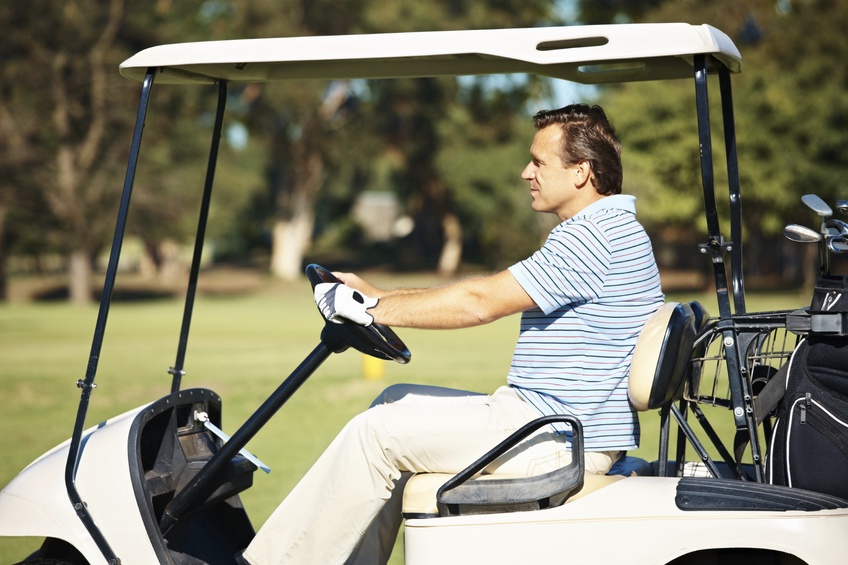 Hombre con una camiseta tipo polo conduciendo un carrito de golf por una calle bordeada de árboles