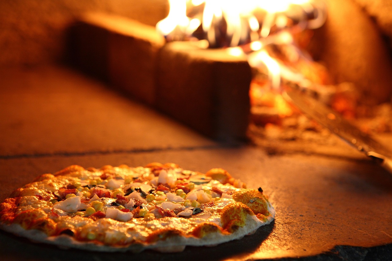 Pizza horneándose sobre la base de piedra de un horno de leña con llamas al fondo