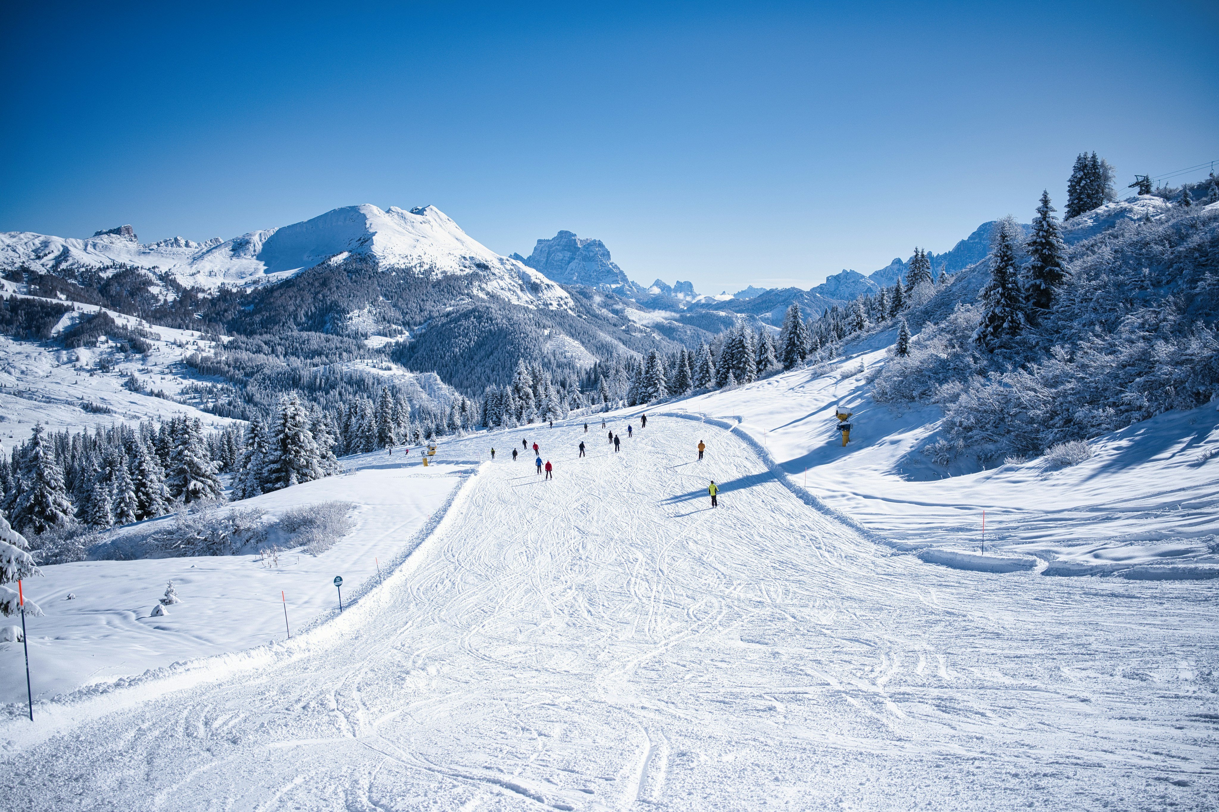 Skiers on a wide groomed run with snow-covered peaks and blue sky