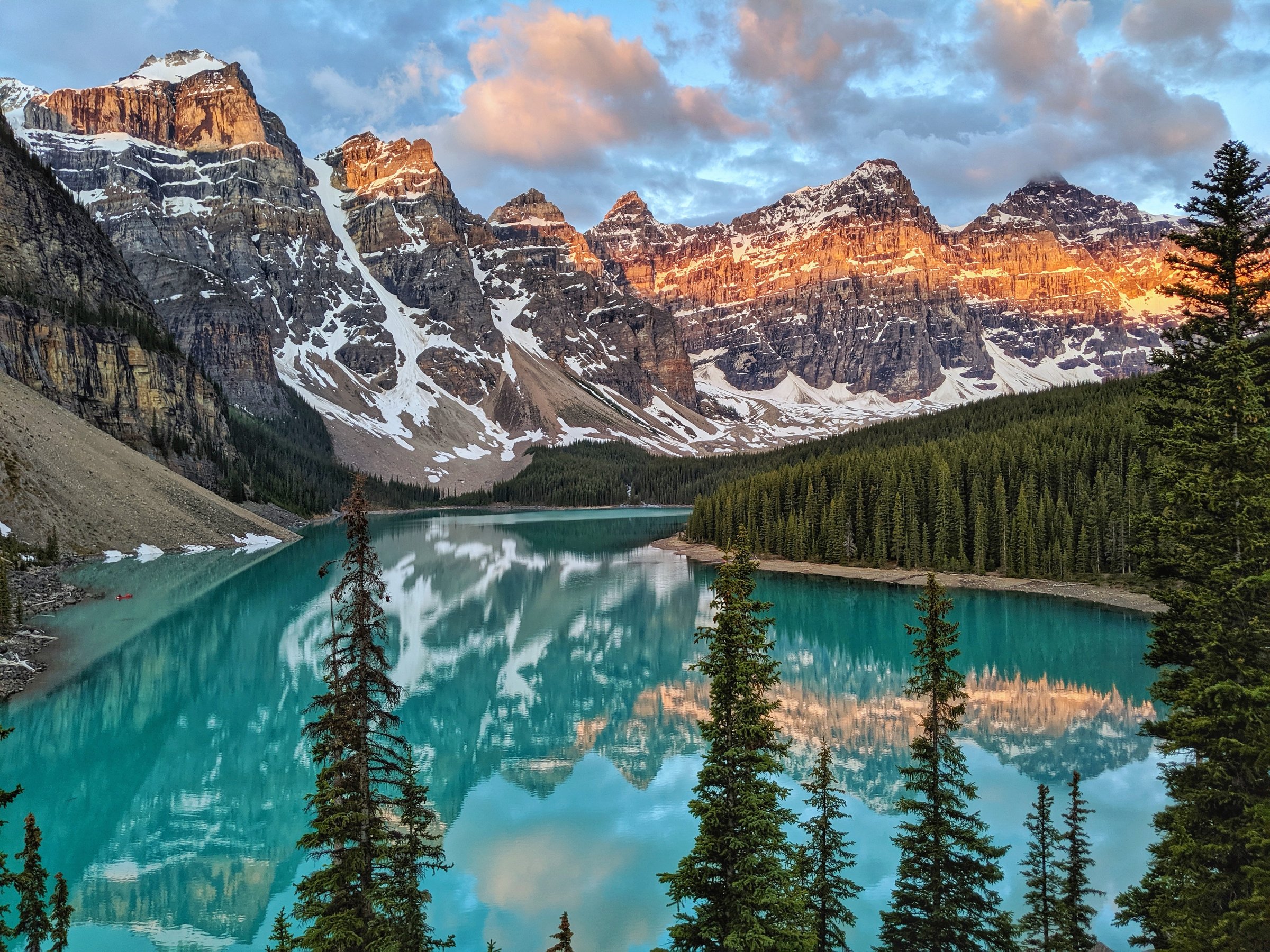 glacial lake surrounded by evergreen trees and large snow capped mountains in the background