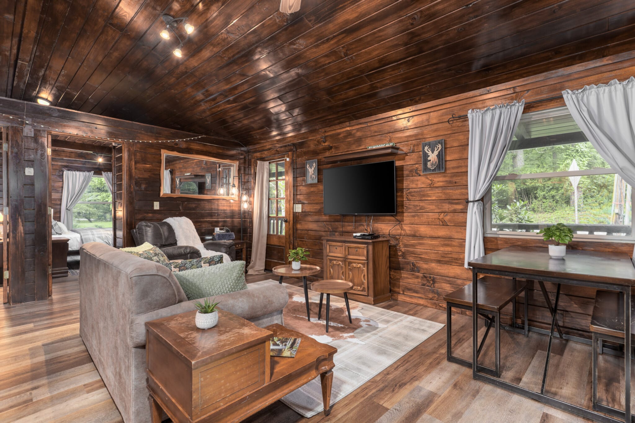 Cozy cabin living room with wooden walls, mounted TV, rustic furniture, and large windows with natural light