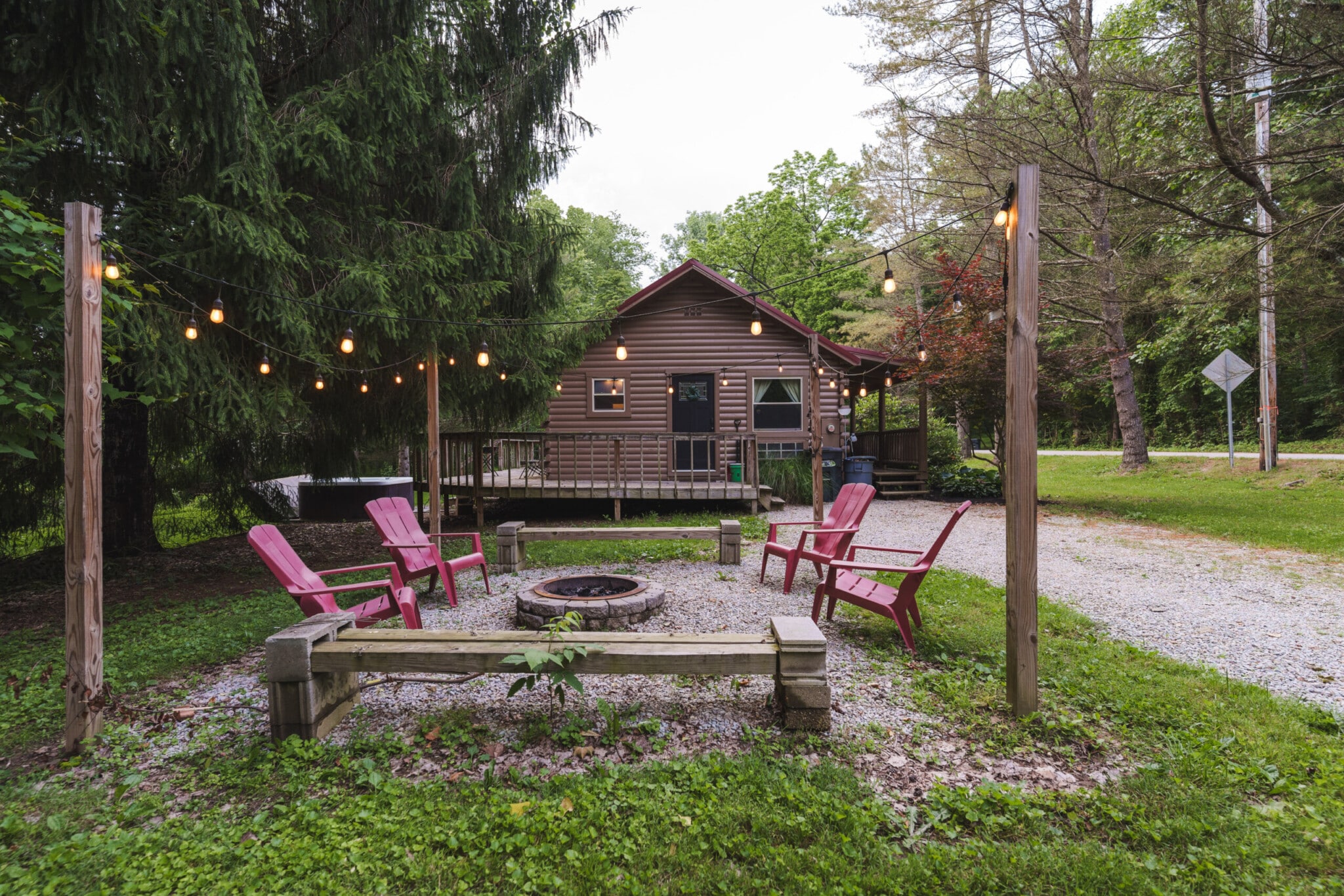 Cozy cabin with string lights, fire pit, and red Adirondack chairs in a wooded backyard
