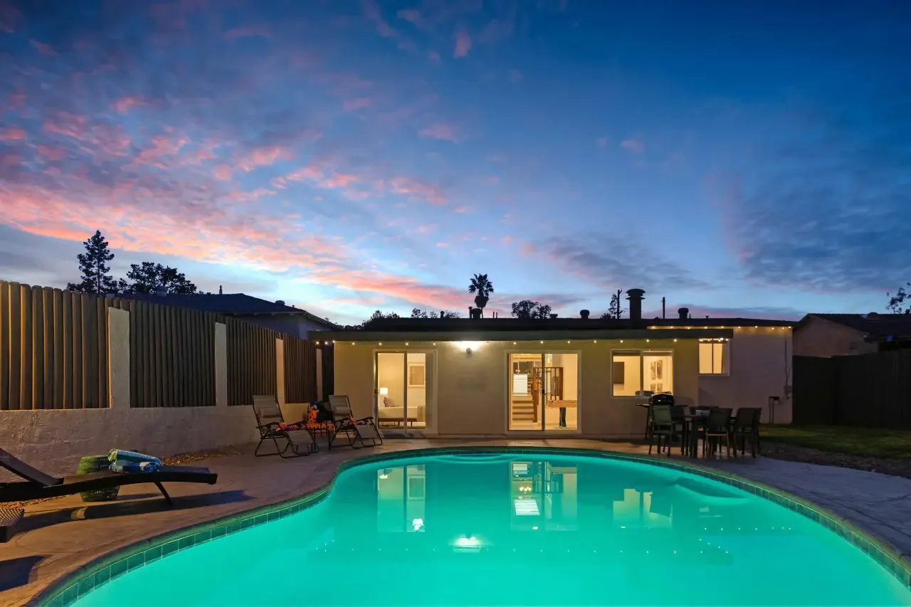 Twilight view of a modern San Diego short-term rental with illuminated turquoise pool, patio seating, and glass doors