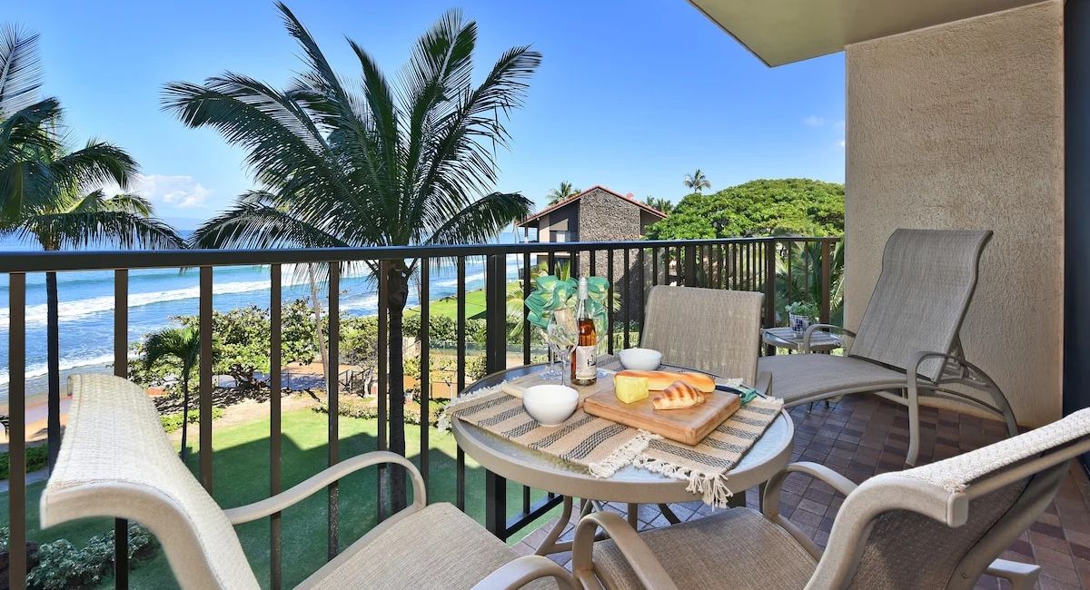 Balcony of Kaanapli Shores 404 overlooking ocean and palm trees, set with chairs and table with cheese, bread, and wine, conveying a serene, tropical ambiance.