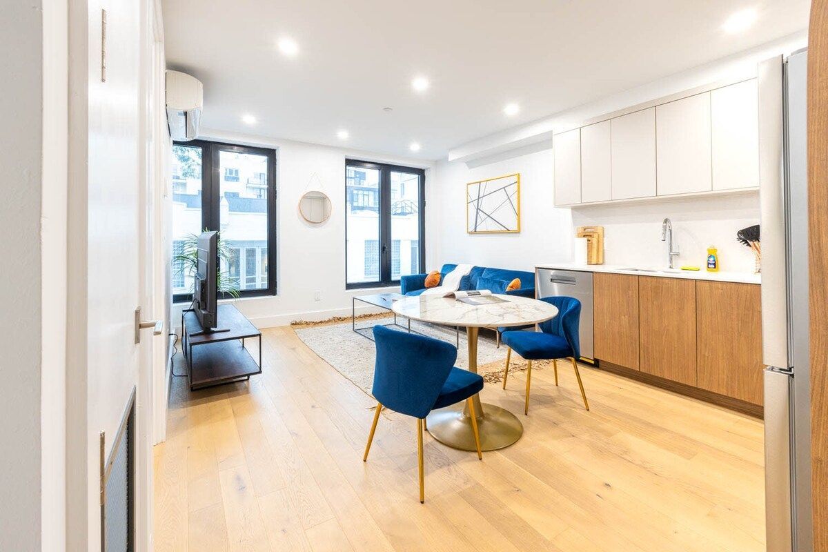 Bright, modern apartment with wood floors. Includes a kitchen, dining area with blue chairs, and a living area with a sofa.