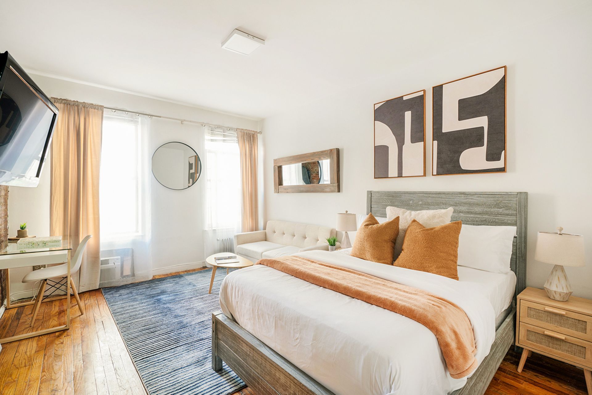 Bedroom with bed, desk, sofa, and art. Neutral tones, blue rug, natural light.