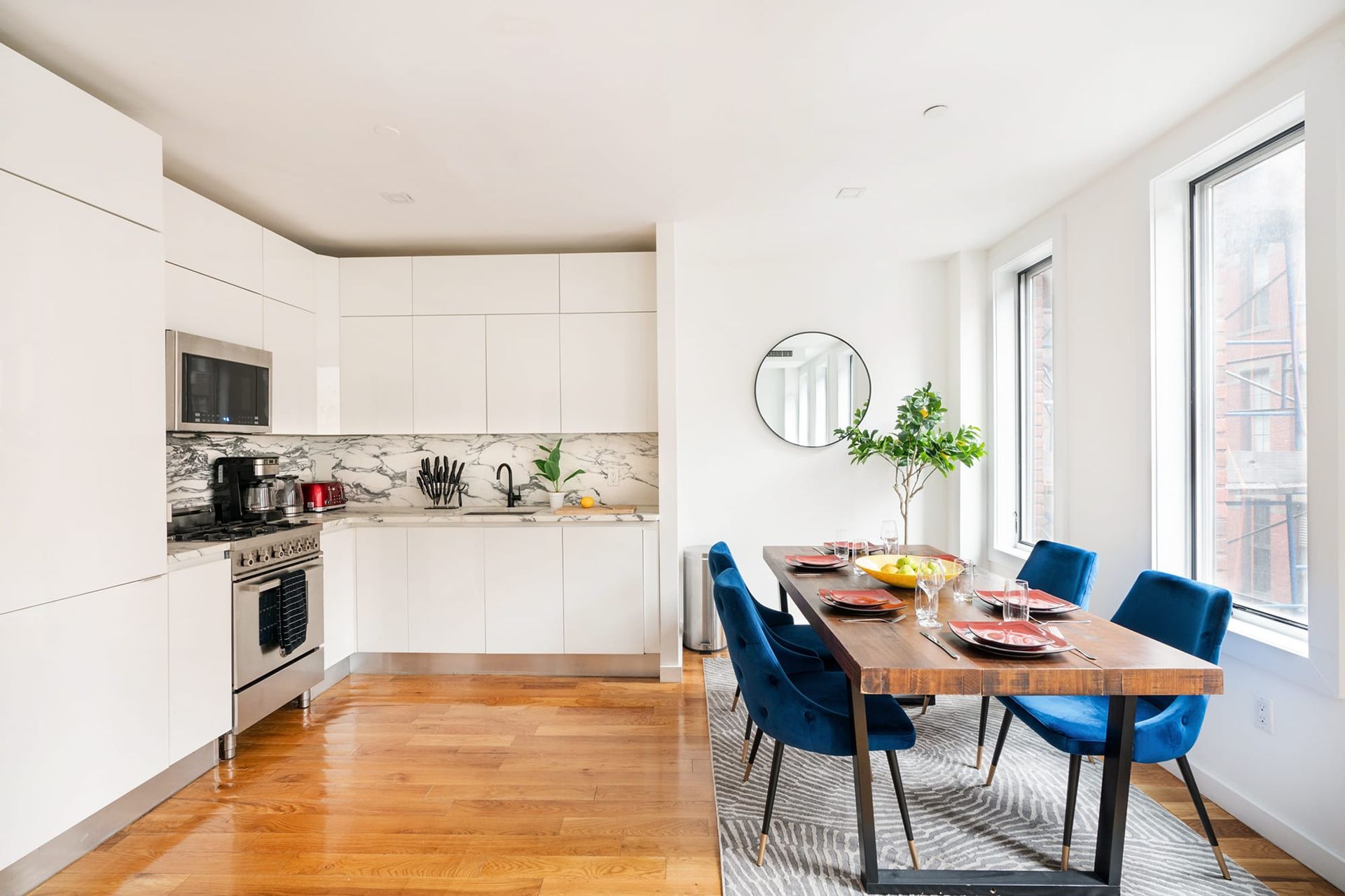 Modern dining room with wood table, blue chairs, white kitchen, and large windows.