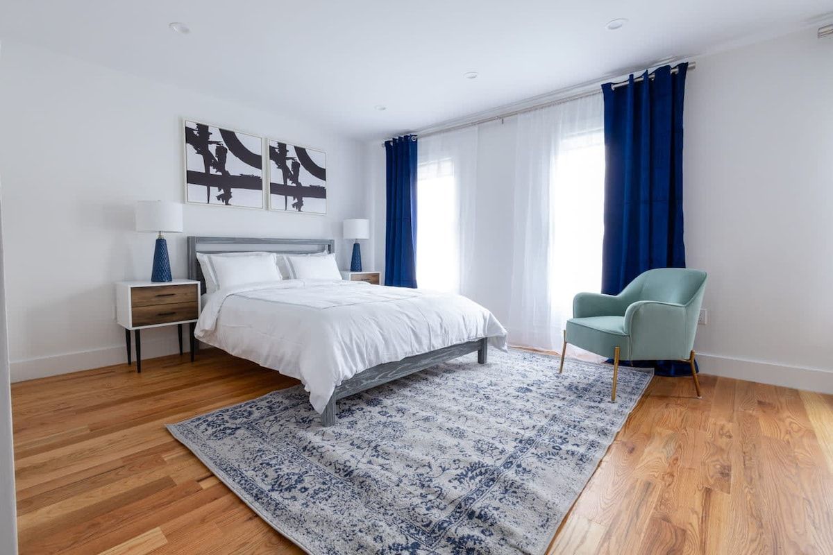 Bedroom with white walls, blue curtains, a bed, armchair, wooden floor, and a patterned rug.