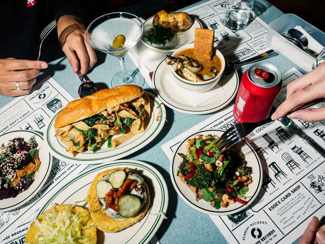 Table with various dishes: sandwich, salad, stew, cocktails, and a soda can. People's hands reach for food.