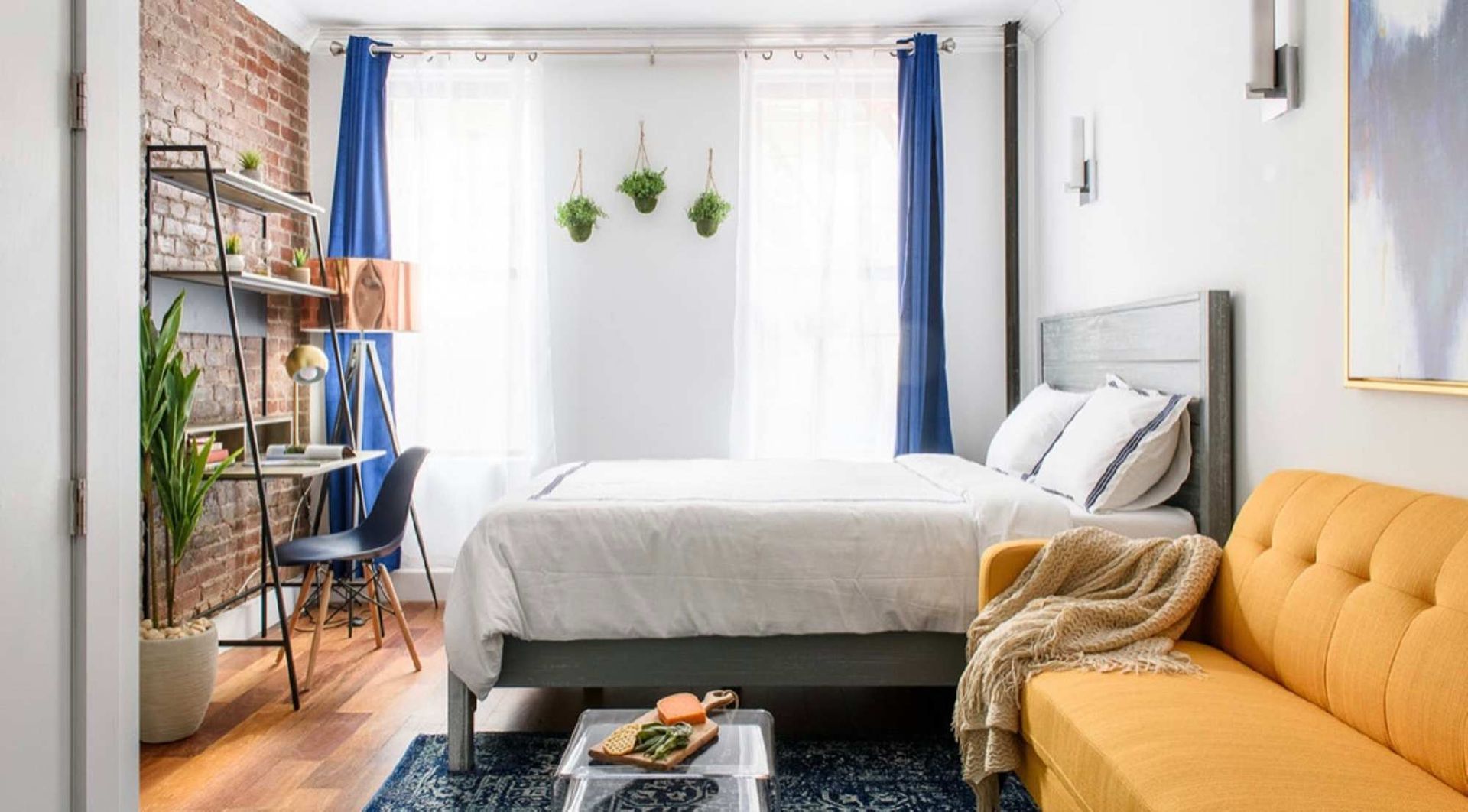 Small bedroom with bed, sofa, desk, and brick wall. Blue and yellow accents.