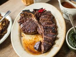 Sliced, cooked T-bone steak on a white plate with sides of sauce and green herbs.