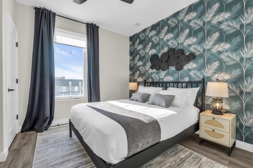 Modern bedroom featuring a queen bed with decorative pillows, tropical wallpaper, and large windows with black curtains, emphasizing coastal-inspired design suitable for shore property rentals.