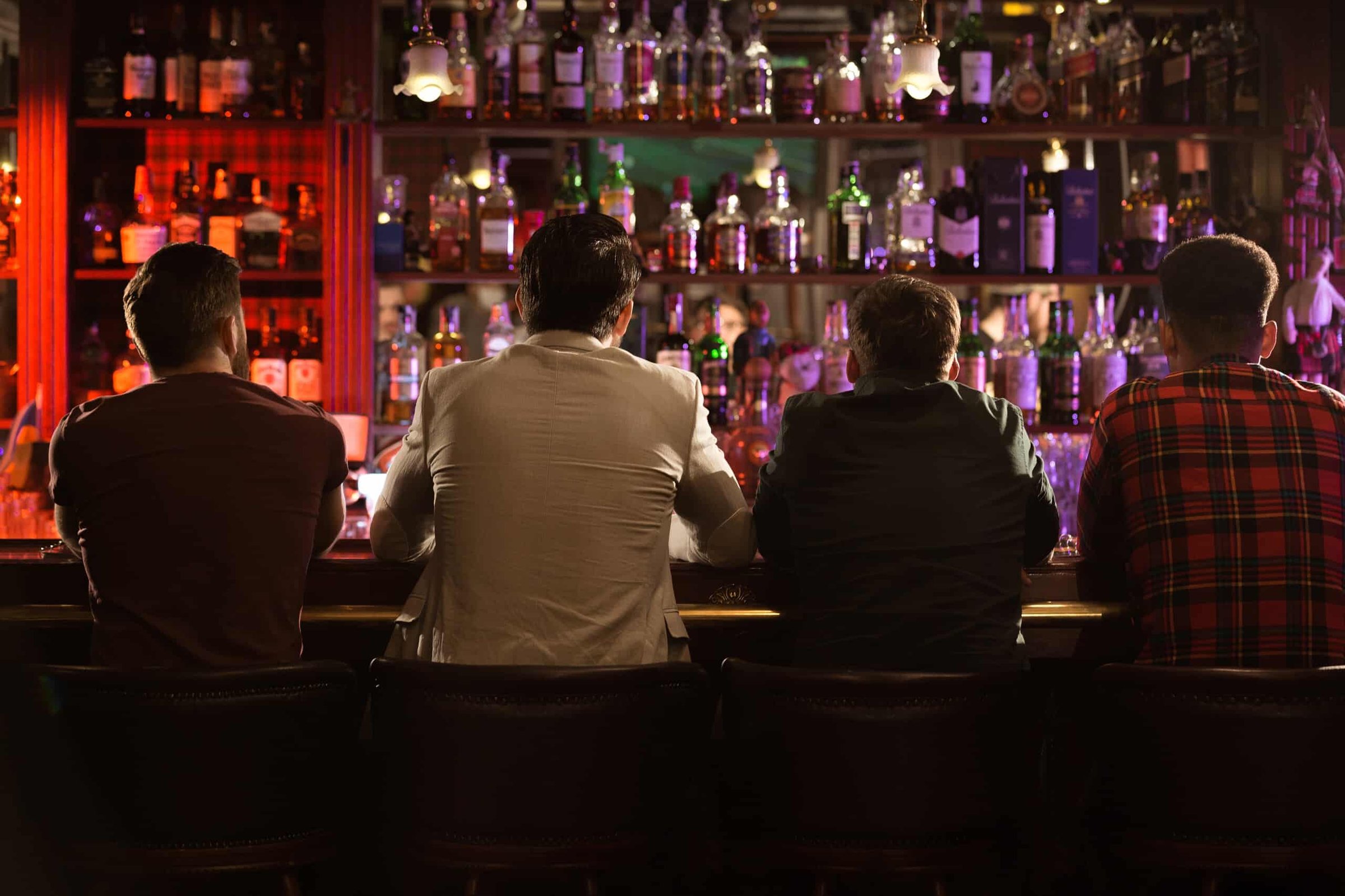 Four young men sitting at a bar, enjoying drinks, with a vibrant display of liquor bottles illuminated in the background, capturing the casual nightlife atmosphere of the Jersey Shore.