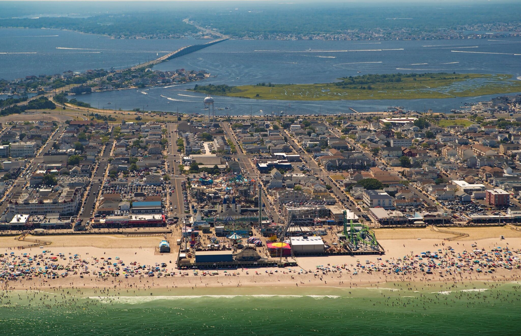 Aerial view of a vibrant Jersey Shore beach town, showcasing sandy beaches filled with umbrellas, a bustling boardwalk with amusement rides, and nearby residential areas, reflecting the lively atmosphere of the local real estate market.