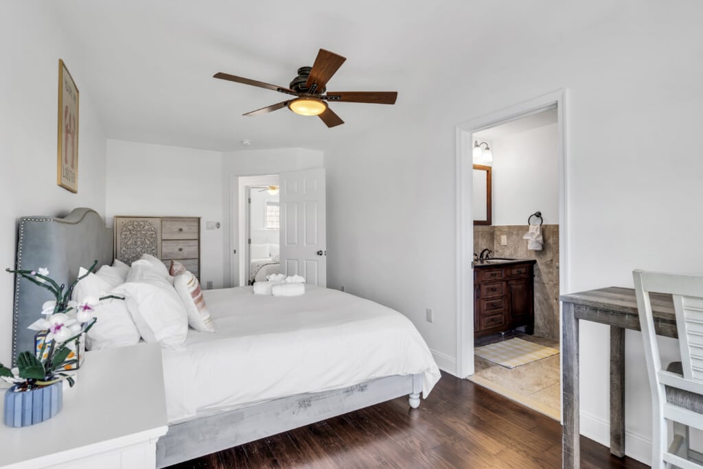 Brightly lit bedroom featuring a king-sized bed with decorative pillows, a wooden ceiling fan, and an en-suite bathroom visible through an open door, emphasizing the cozy and inviting atmosphere ideal for vacation rentals in Manasquan.