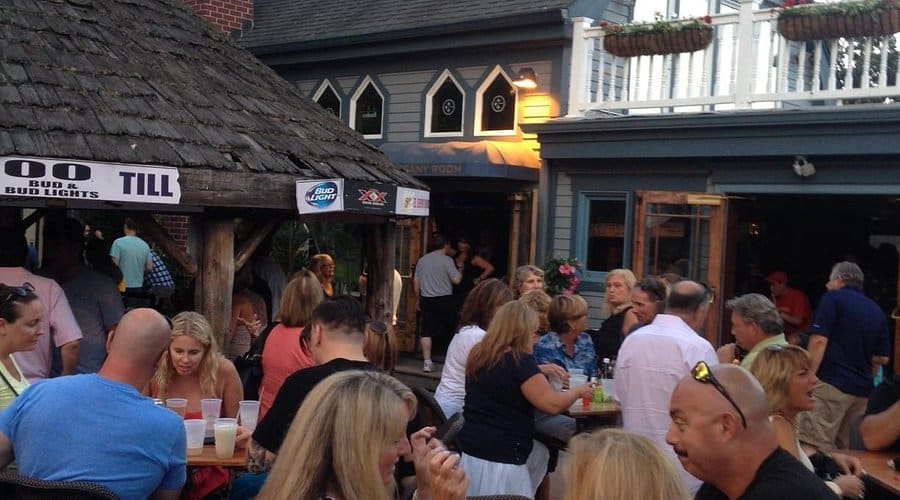 Lively outdoor bar scene at a Jersey Shore venue, featuring patrons enjoying drinks and socializing under a thatched roof, with a bustling atmosphere and bar signage visible.