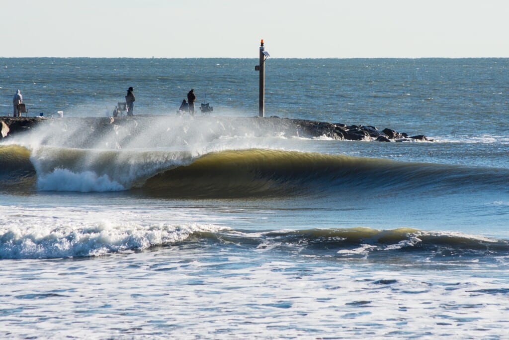 Surf wave breaking near jetty at Nun’s Beach, with surfers and beachgoers in the background, showcasing ideal conditions for beginner and intermediate surfers.