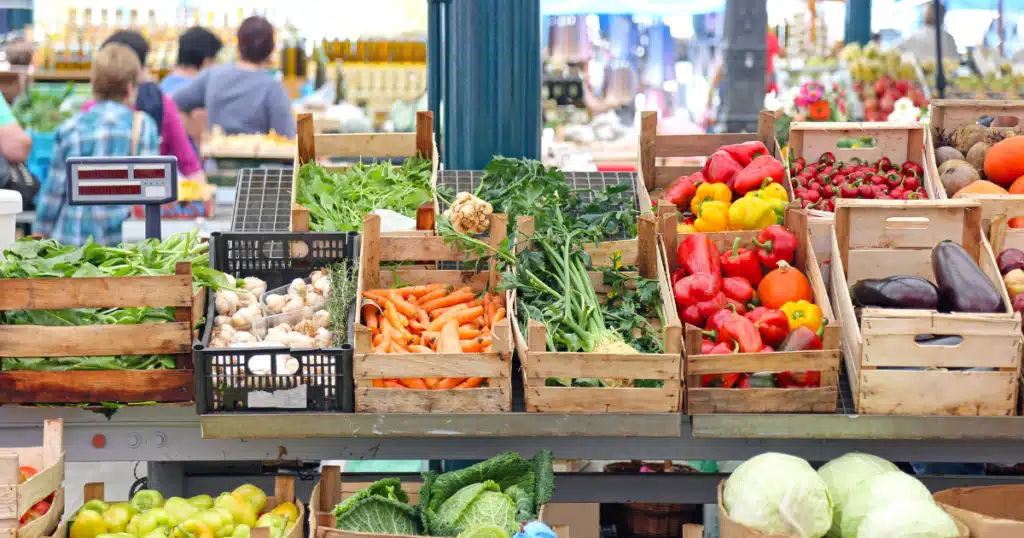 Farmers market display featuring fresh vegetables and fruits, including carrots, peppers, garlic, and leafy greens, with shoppers in the background, capturing the vibrant community atmosphere at the Jersey Shore.