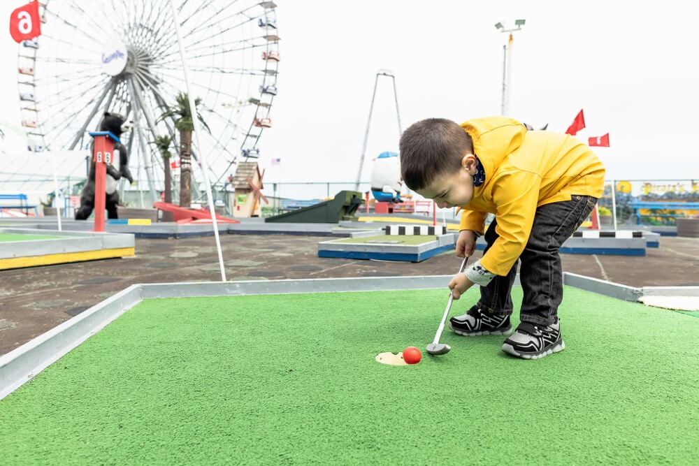 Child in yellow jacket playing miniature golf on a vibrant green course, with a ferris wheel and colorful obstacles in the background, capturing the fun and excitement of family-friendly activities at Ortley Beach's pirate-themed mini golf.
