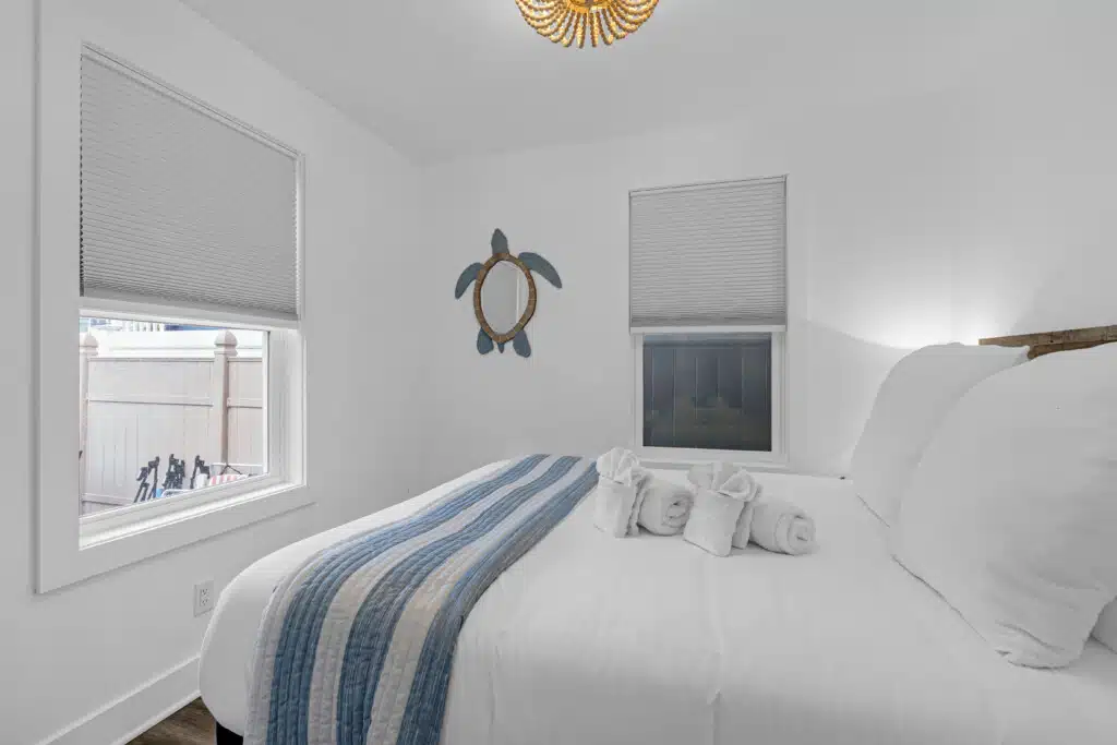 Cozy bedroom featuring a queen bed with striped bedding, decorative turtle wall art, and natural light from two windows, ideal for family-friendly beach stays at the Jersey Shore.
