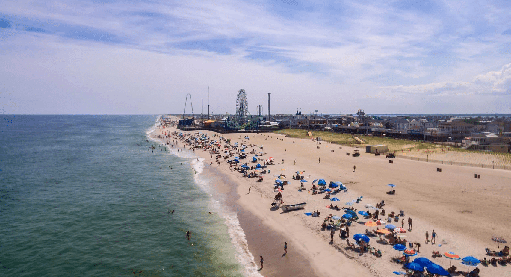 Aerial view of a busy Jersey Shore beach with colorful umbrellas, people enjoying the sun, and amusement rides in the background, highlighting the vibrant atmosphere of summer vacations.