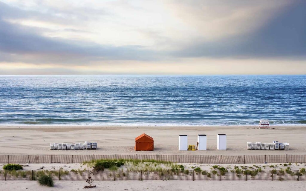 Beachfront view at Jersey Shore featuring orange cabana, white beach huts, and rows of lounge chairs along pristine sandy shore, with calm ocean waves under a cloudy sky.
