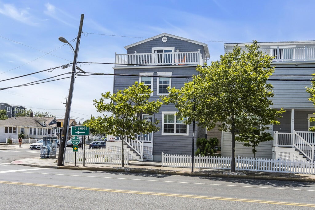 Gray beach house with balconies, surrounded by trees and a white picket fence, near street sign directing to the bridge, indicative of vacation rental options at the Jersey Shore.
