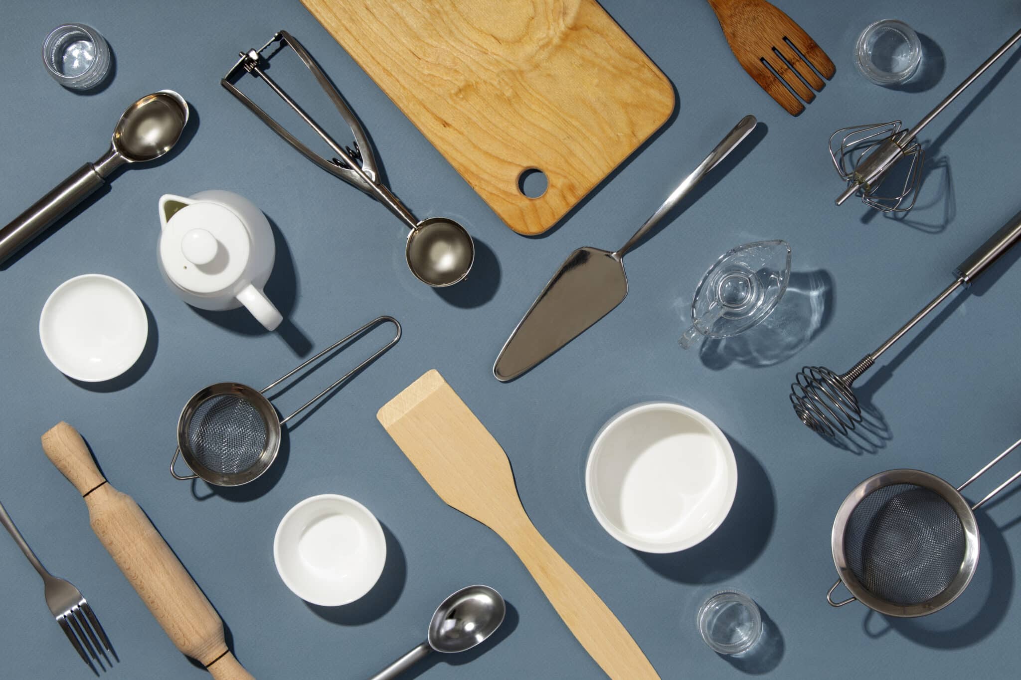 Kitchen utensils and supplies arranged on a blue background, including a wooden cutting board, measuring cups, a teapot, a whisk, and various cooking tools, reflecting essential items for preparing meals during a beach vacation.