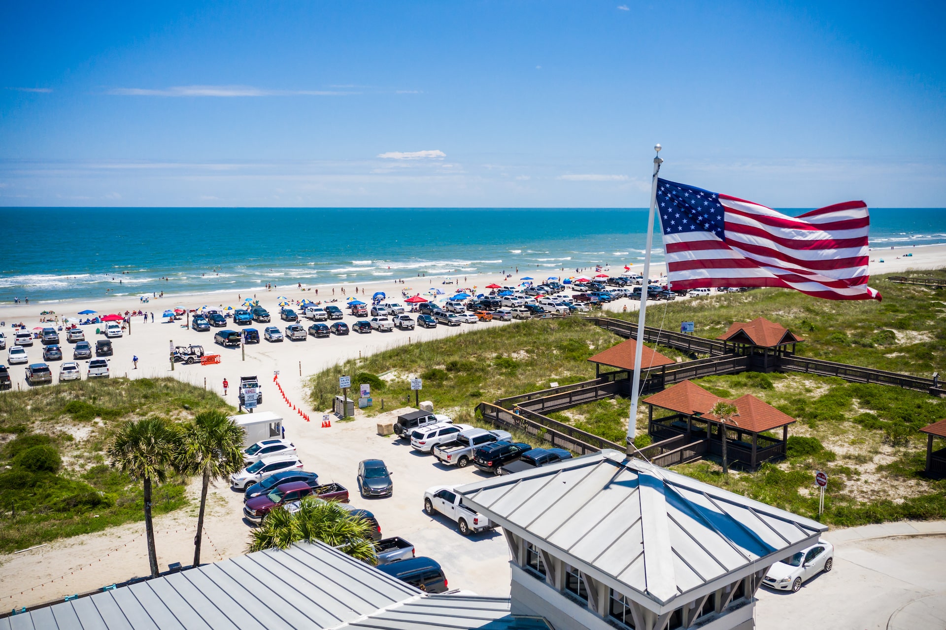 Aerial view of Seaside Park beach with cars parked near the shore, colorful umbrellas, and an American flag waving in the foreground, highlighting beach access and amenities for vacationers.