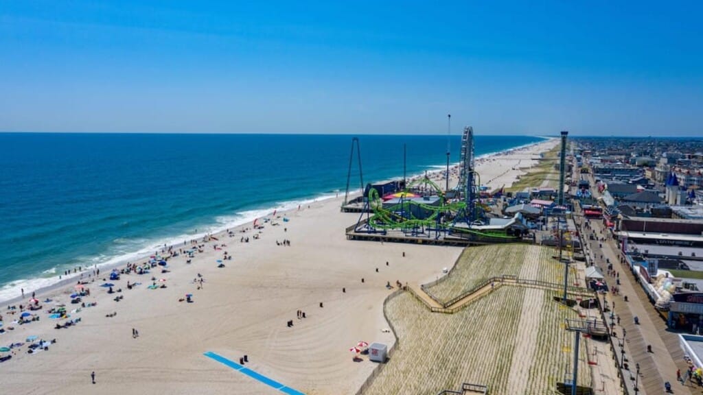 Aerial view of Seaside Heights beach with families enjoying the sun, amusement park rides in the background, and a vibrant boardwalk lined with shops and attractions.