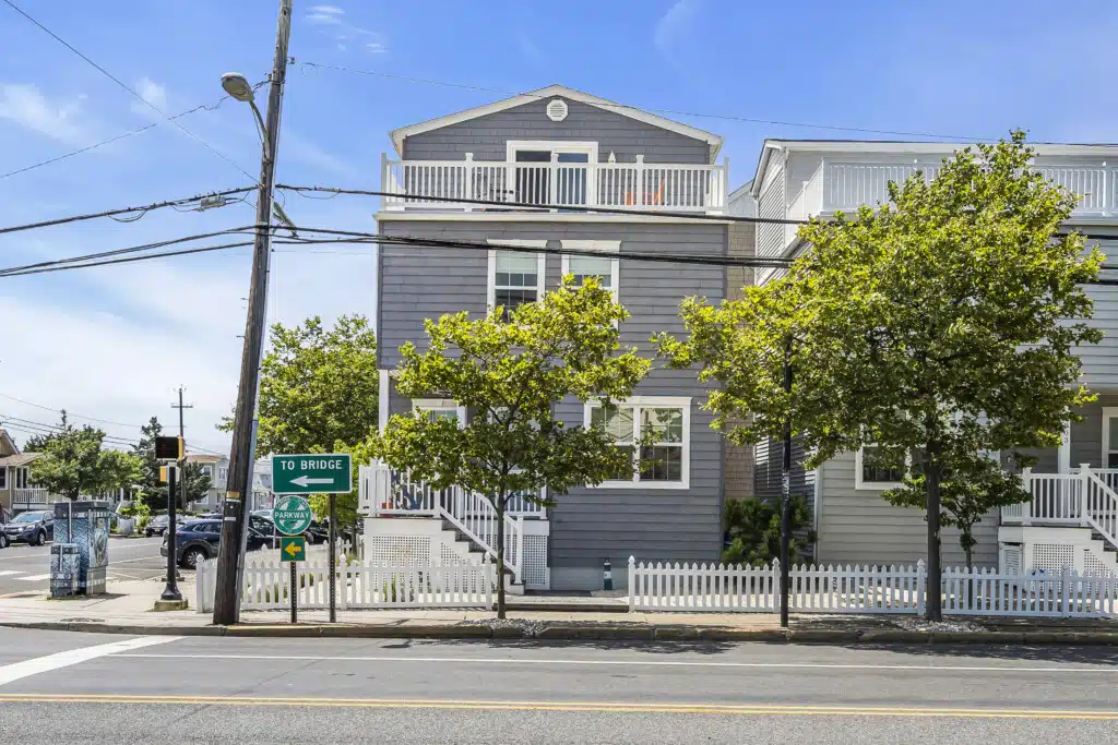 Coastal home exterior with gray siding, white trim, and balconies, surrounded by greenery and street signs indicating directions to the bridge and parkway, reflecting Jersey Shore property appeal.