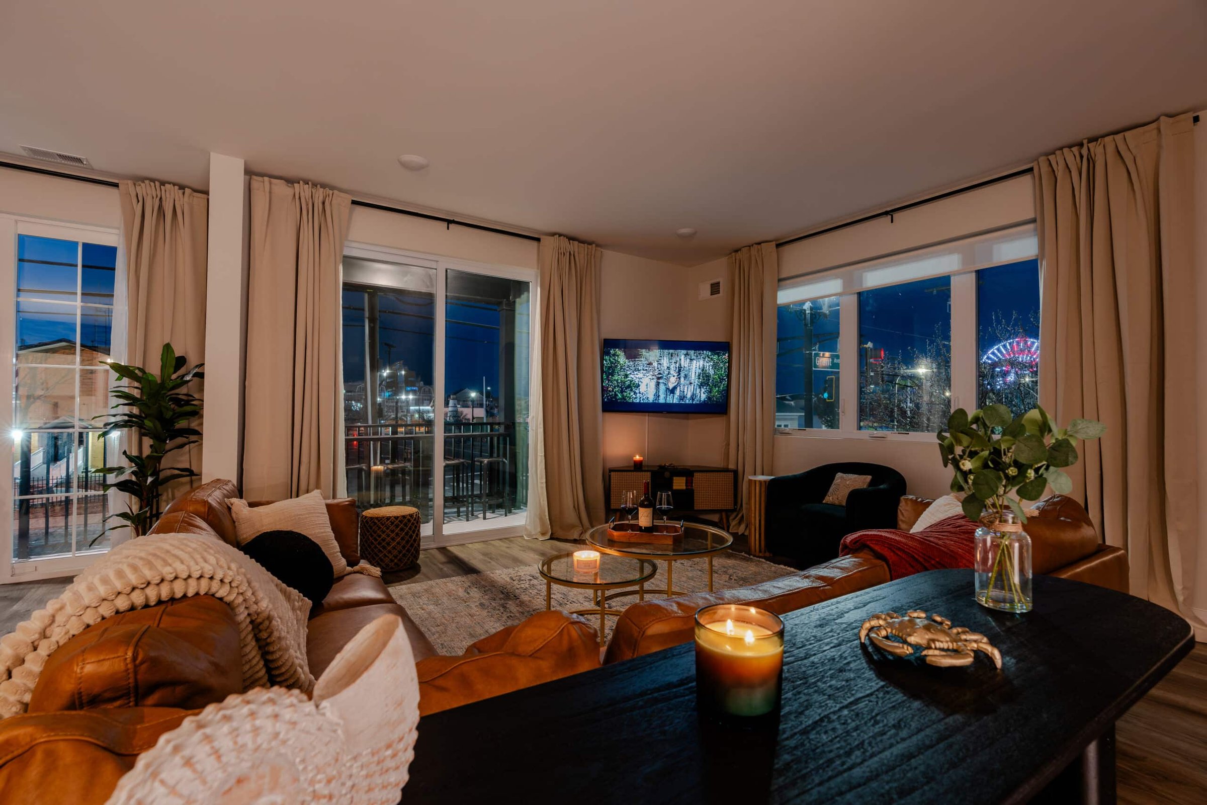 Cozy living room with leather sofa, decorative pillows, and a view of the Jersey Shore skyline at night, featuring a TV, coffee table with candles, and potted plant, suitable for vacation rentals.