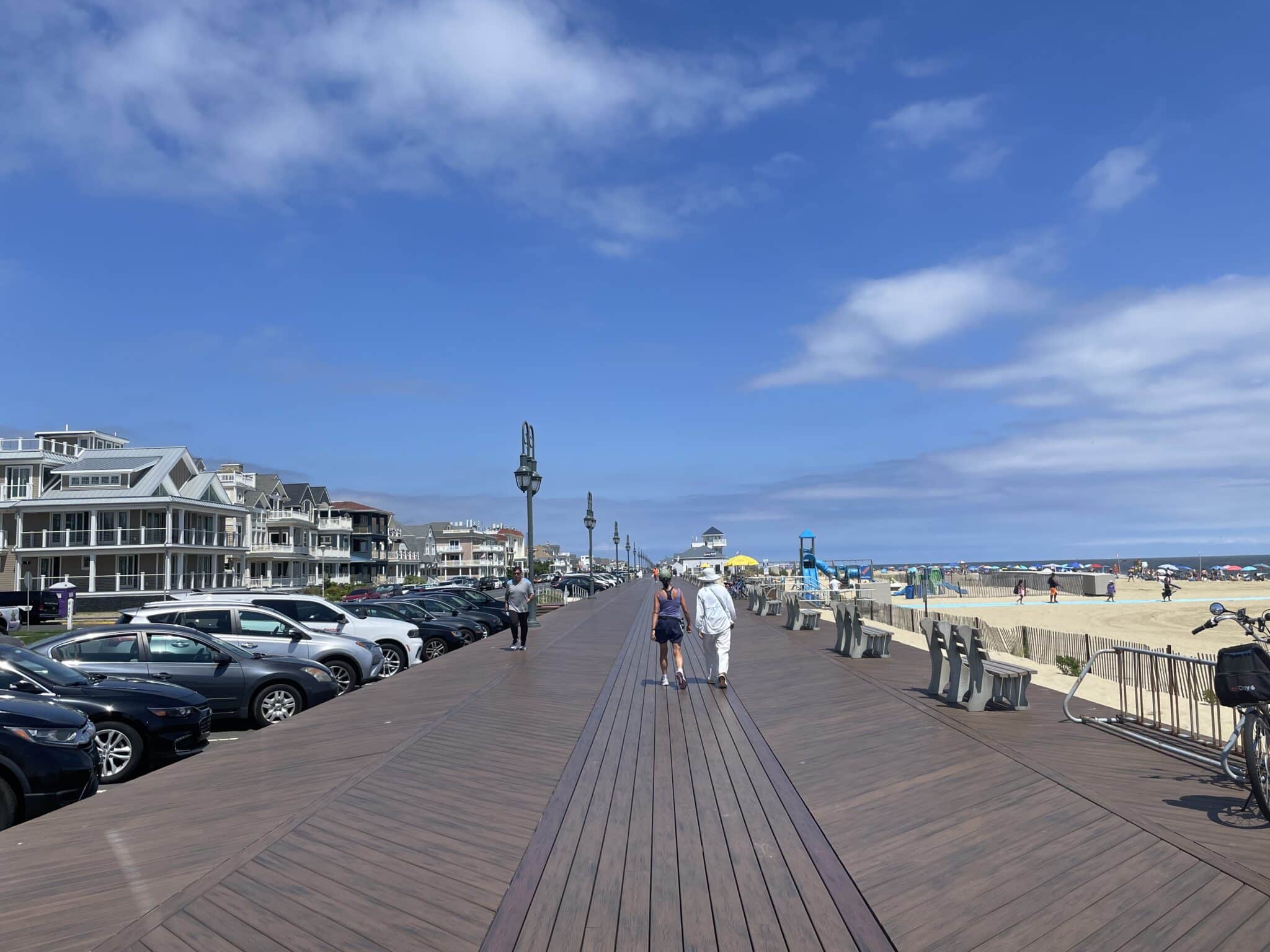 Belmar boardwalk view with pedestrians walking, beach and ocean in the background, nearby parking, and beach amenities visible.