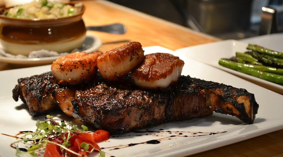 Grilled steak topped with seared scallops, garnished with microgreens and cherry tomatoes, accompanied by asparagus and a bowl of fried rice, representing family-friendly dining options at Breezy Beach Stays.