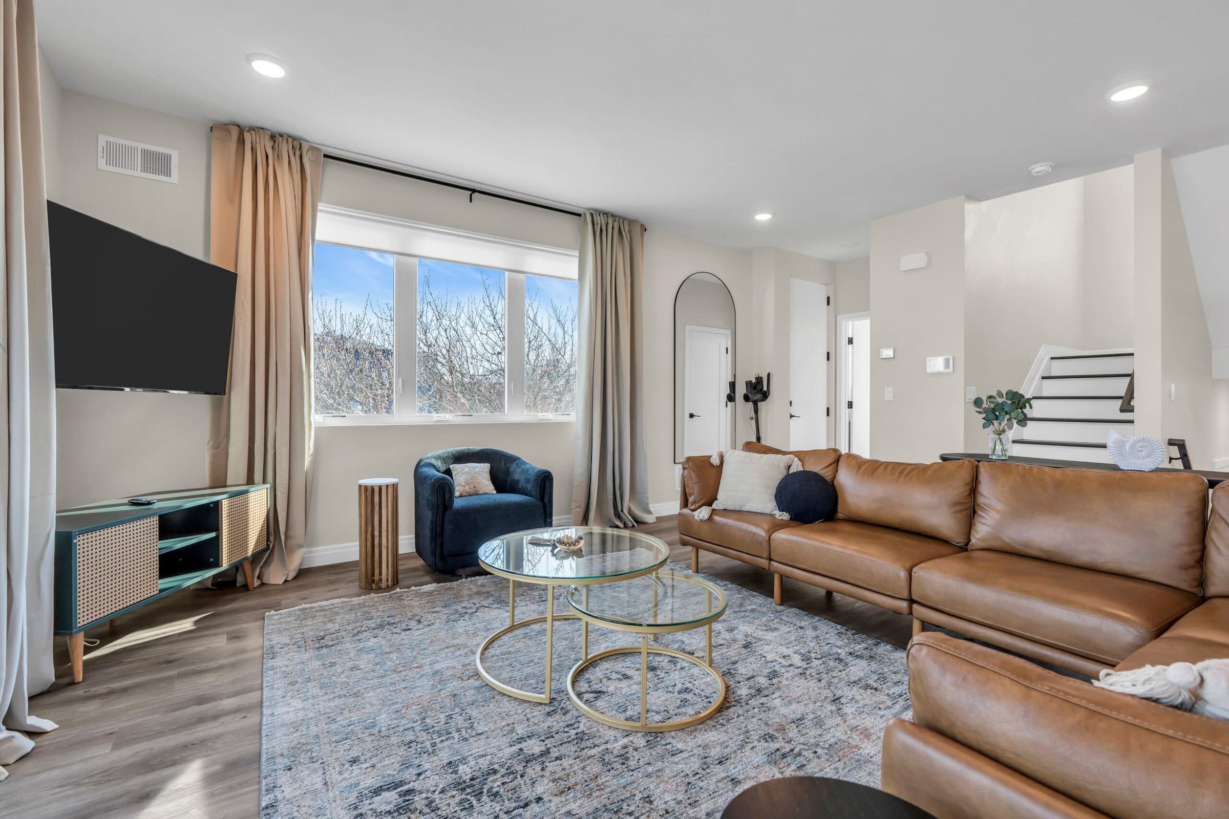 Modern living room featuring a brown leather sectional sofa, glass coffee tables, and a blue accent chair, with large windows allowing natural light and a view of trees outside, ideal for vacation rentals at the Jersey Shore.