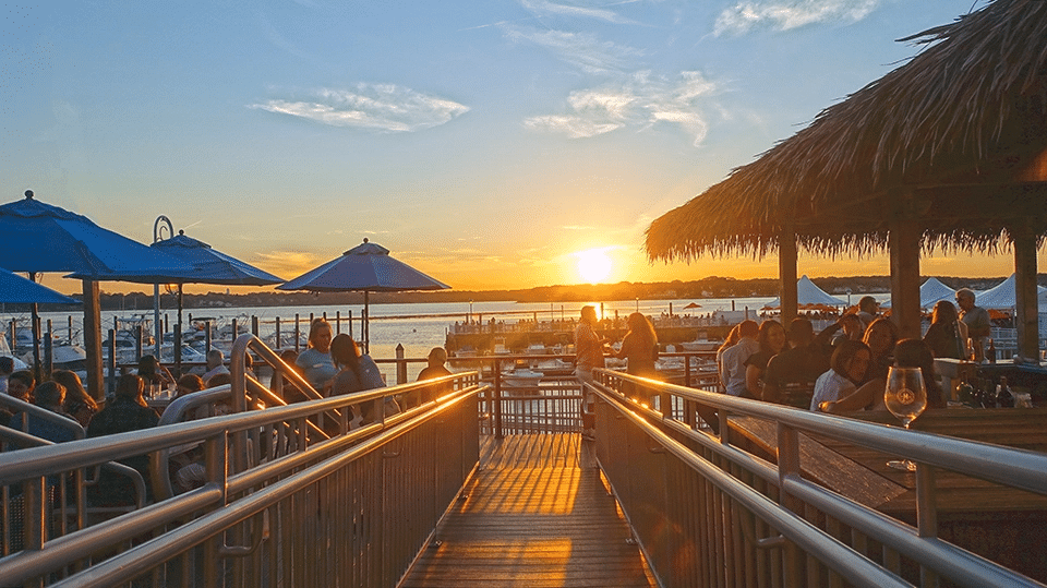 Outdoor dining at sunset in Belmar, featuring patrons enjoying ocean views, blue umbrellas, and a vibrant atmosphere near the Jersey Shore.