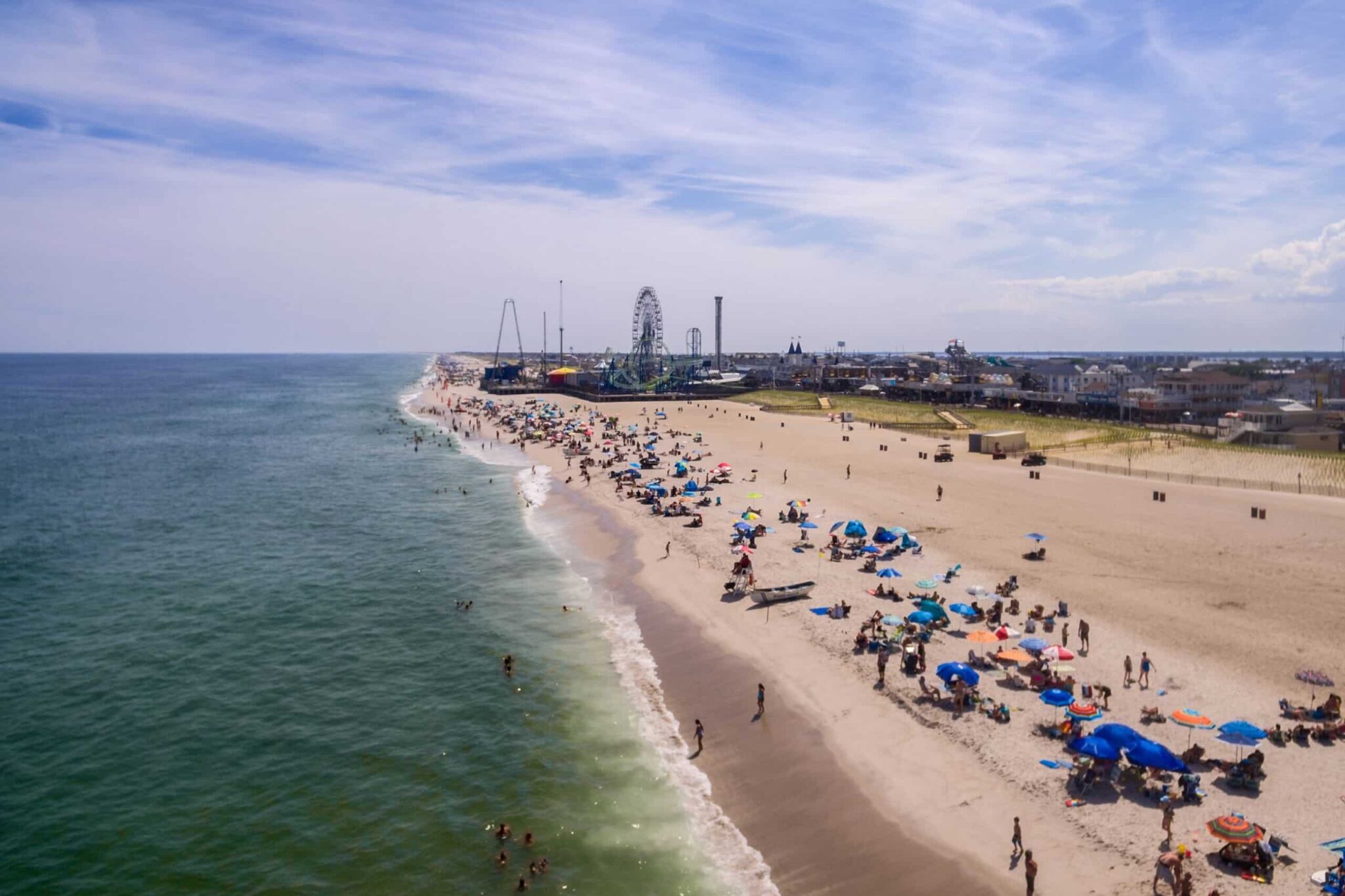 Aerial view of Seaside Heights beach with colorful umbrellas, people enjoying the sun, and the iconic boardwalk featuring amusement rides in the background.