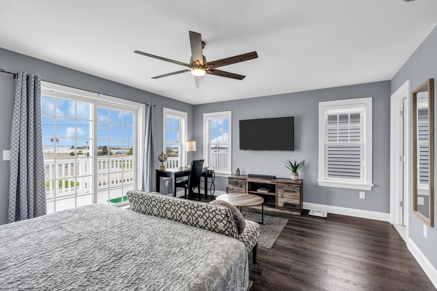 Modern vacation rental interior in Seaside Heights, featuring a spacious bedroom with a patterned bedspread, a desk, a TV, large windows with natural light, and coastal decor, highlighting Breezy Beach Stays' management success.