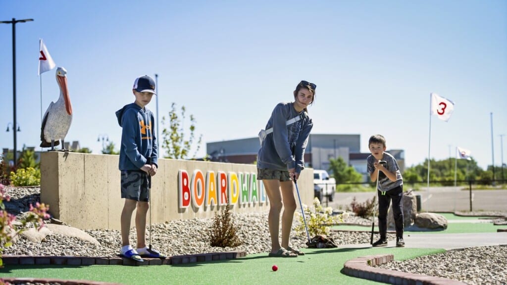 Family-friendly mini-golf attraction at the Jersey Shore, featuring children and a young adult playing, with a colorful "BOARDWALK" sign and a pelican statue in the background.