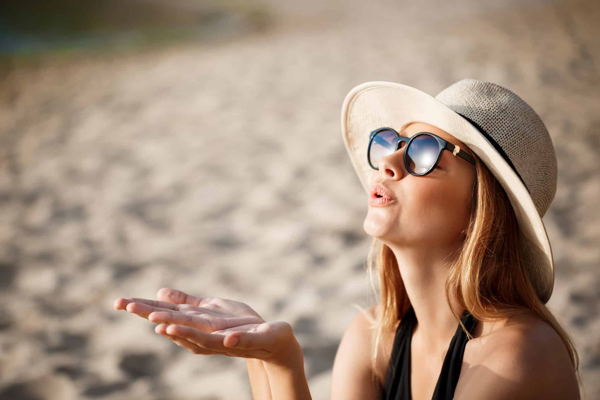Young woman wearing a wide-brimmed sun hat and sunglasses, enjoying a sunny day at the beach, with sand in the background, illustrating essential beach accessories for a vacation.