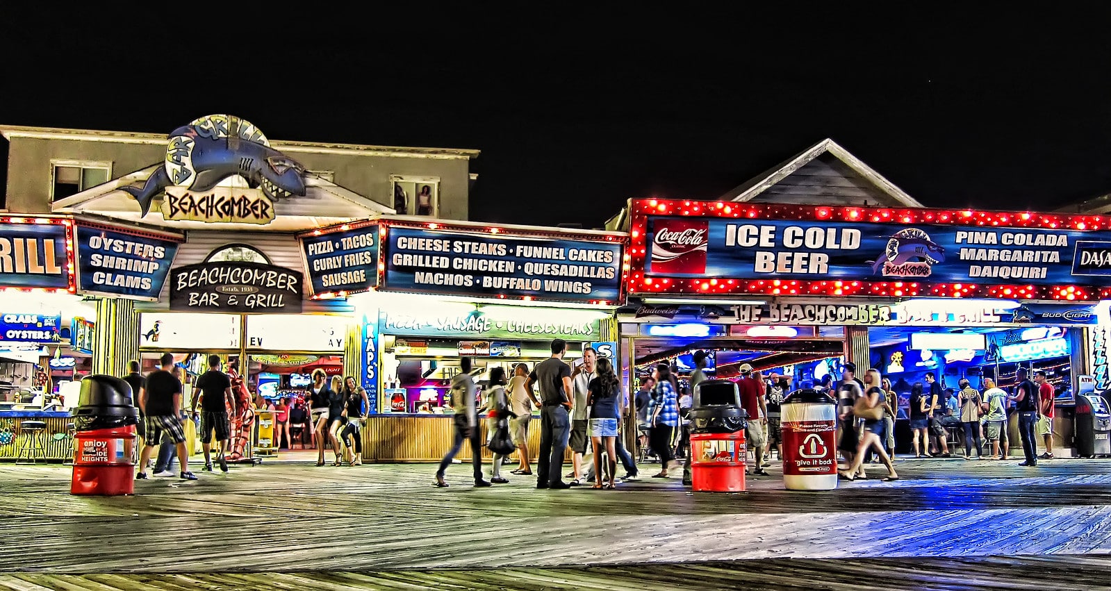 Nighttime scene at Seaside Heights Boardwalk featuring the Beachcomber Bar & Grill, showcasing vibrant signage for seafood, drinks, and live entertainment, with a lively crowd enjoying the nightlife atmosphere.