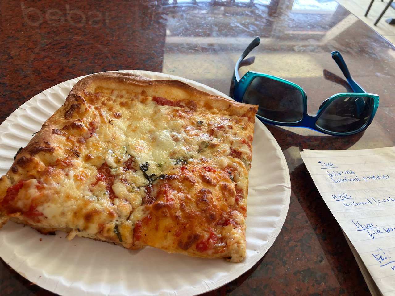 Slice of thin, crispy boardwalk pizza with melted cheese and toppings on a paper plate, accompanied by blue sunglasses and a notepad, capturing the casual dining experience at the Jersey Shore.